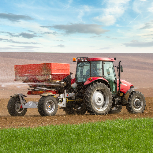 Red tractor fertilizing an empty field. In the foreground, the field is lined with grass. The background features a blue sky dotted with scattered grey and white clouds.